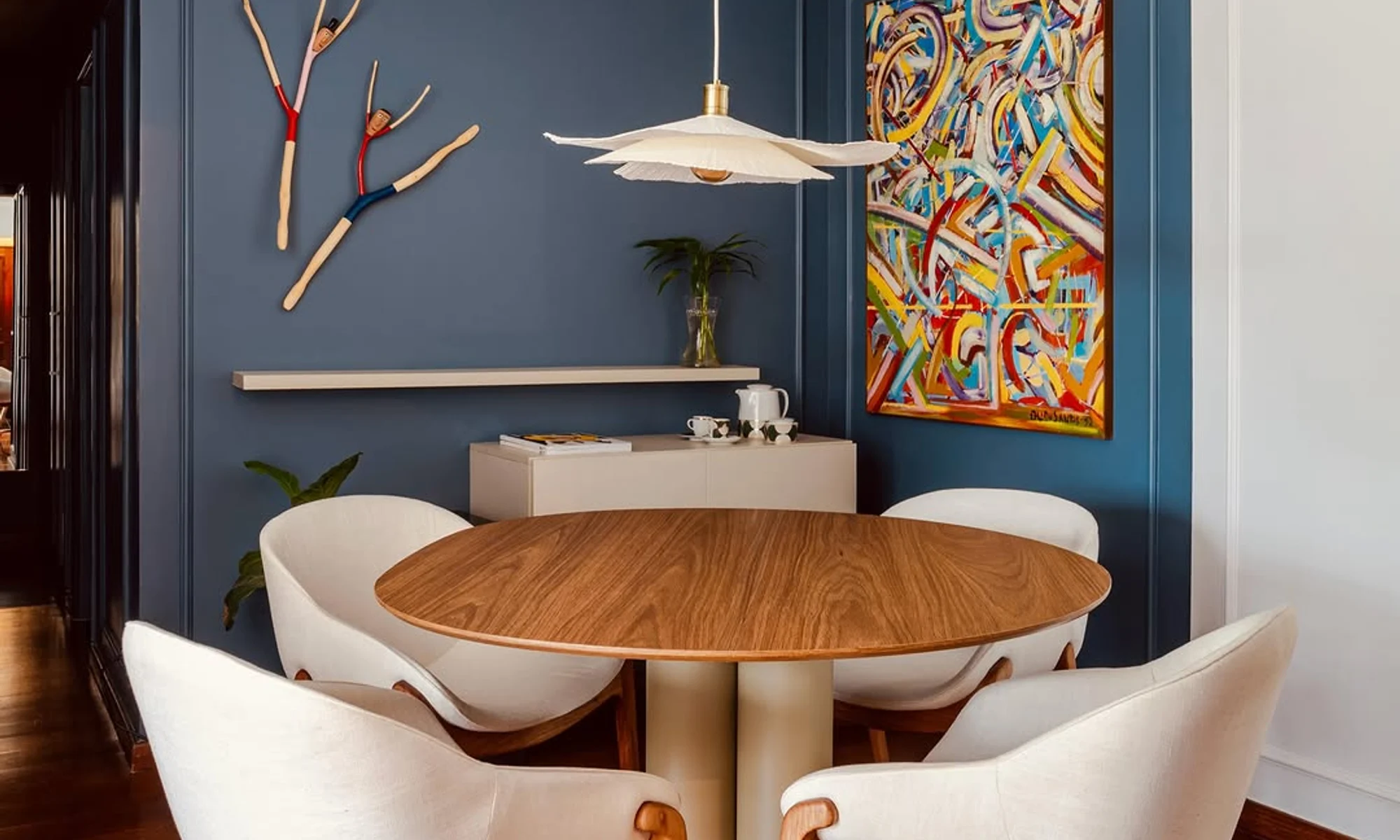 Sala de jantar com parede azul petróleo, arte colorida, mesa redonda de madeira e cadeiras escultóricas, projeto da Um pra Um